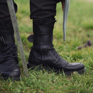 Op de afbeelding: Zwarte leren laarzen met een vetersluiting en dikke zolen. De laarzen hebben een band en gesp detail rond de enkel. De laarzen worden gedragen met een zwarte broek en staan tegen een achtergrond van groen gras.