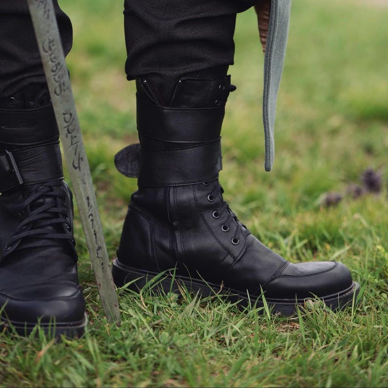 Op de afbeelding: Zwarte leren laarzen met een vetersluiting en dikke zolen. De laarzen hebben een band en gesp detail rond de enkel. De laarzen worden gedragen met een zwarte broek en staan tegen een achtergrond van groen gras.