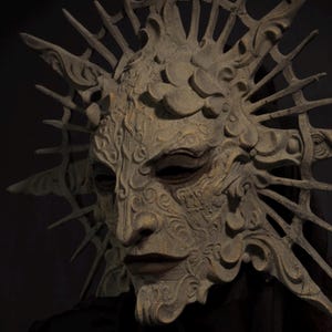 May include: A detailed, antique-style mask with a textured, weathered appearance. The mask features intricate carvings, a prominent nose, and a crown-like structure with sharp, radiating points. The color is a muted, earthy tone, suggesting stone or aged material. The mask is set against a dark background.
