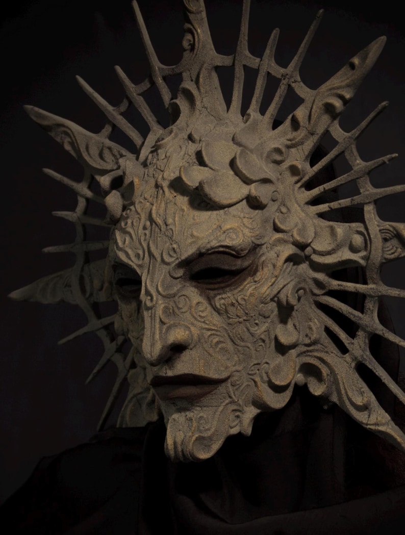 May include: A detailed, antique-style mask with a textured, weathered appearance. The mask features intricate carvings, a prominent nose, and a crown-like structure with sharp, radiating points. The color is a muted, earthy tone, suggesting stone or aged material. The mask is set against a dark background.