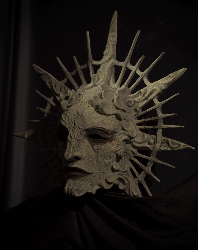 May include: A detailed, off-white mask with a sunburst design. The mask features intricate carvings and a serene facial expression. The mask is set against a dark background, highlighting its ornate details and texture.