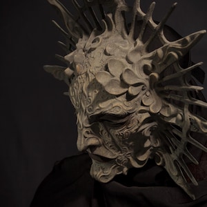 May include: A detailed, ornate mask with a weathered, stone-like appearance. The mask features intricate carvings, a prominent brow, and a crown-like structure with sharp, spiky elements. The overall design suggests a fantasy or theatrical theme.