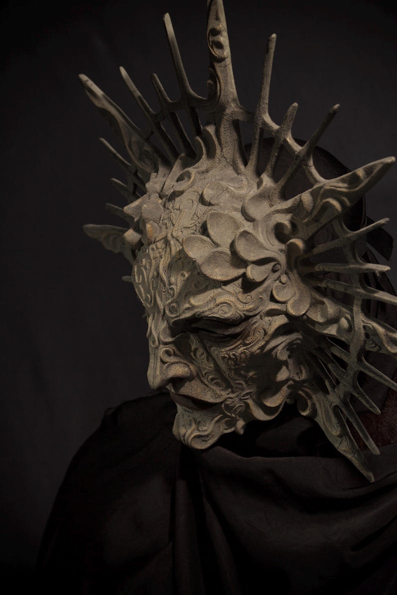 May include: A detailed, ornate mask with a weathered, stone-like appearance. The mask features intricate carvings, a prominent brow, and a crown-like structure with sharp, spiky elements. The overall design suggests a fantasy or theatrical theme.