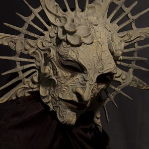 May include: A detailed, weathered mask with an elaborate design. The mask is primarily beige with darker shading and features intricate carvings, a prominent nose, and a sunburst-like crown. The mask is set against a dark background, highlighting its texture and form.