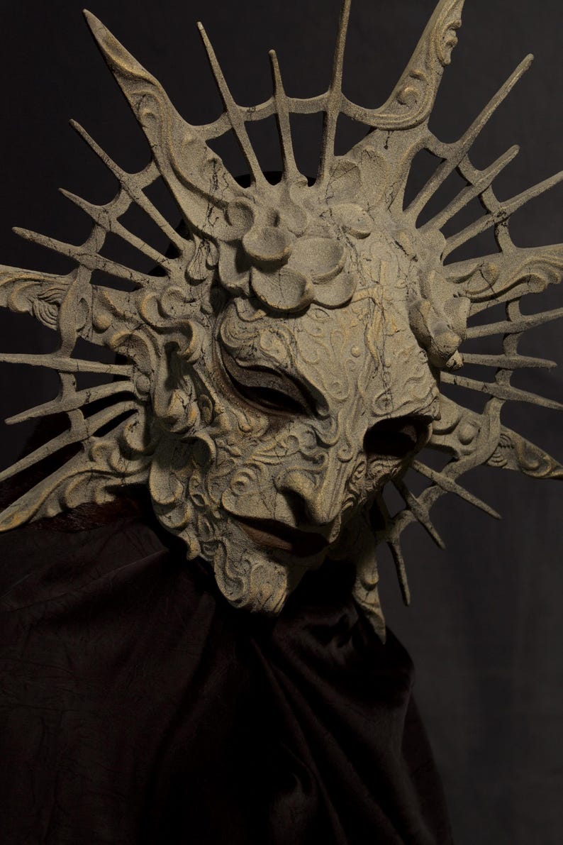 May include: A detailed, weathered mask with an elaborate design. The mask is primarily beige with darker shading and features intricate carvings, a prominent nose, and a sunburst-like crown. The mask is set against a dark background, highlighting its texture and form.