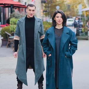 Op de afbeelding: Twee mensen in lange, teal-kleurige jassen. De jas links heeft beige mouwen en wordt gecombineerd met een zwarte broek en bruine laarzen. De jas rechts is donkerder teal en heeft een bijpassende riem. De achtergrond is een stadsstraat.