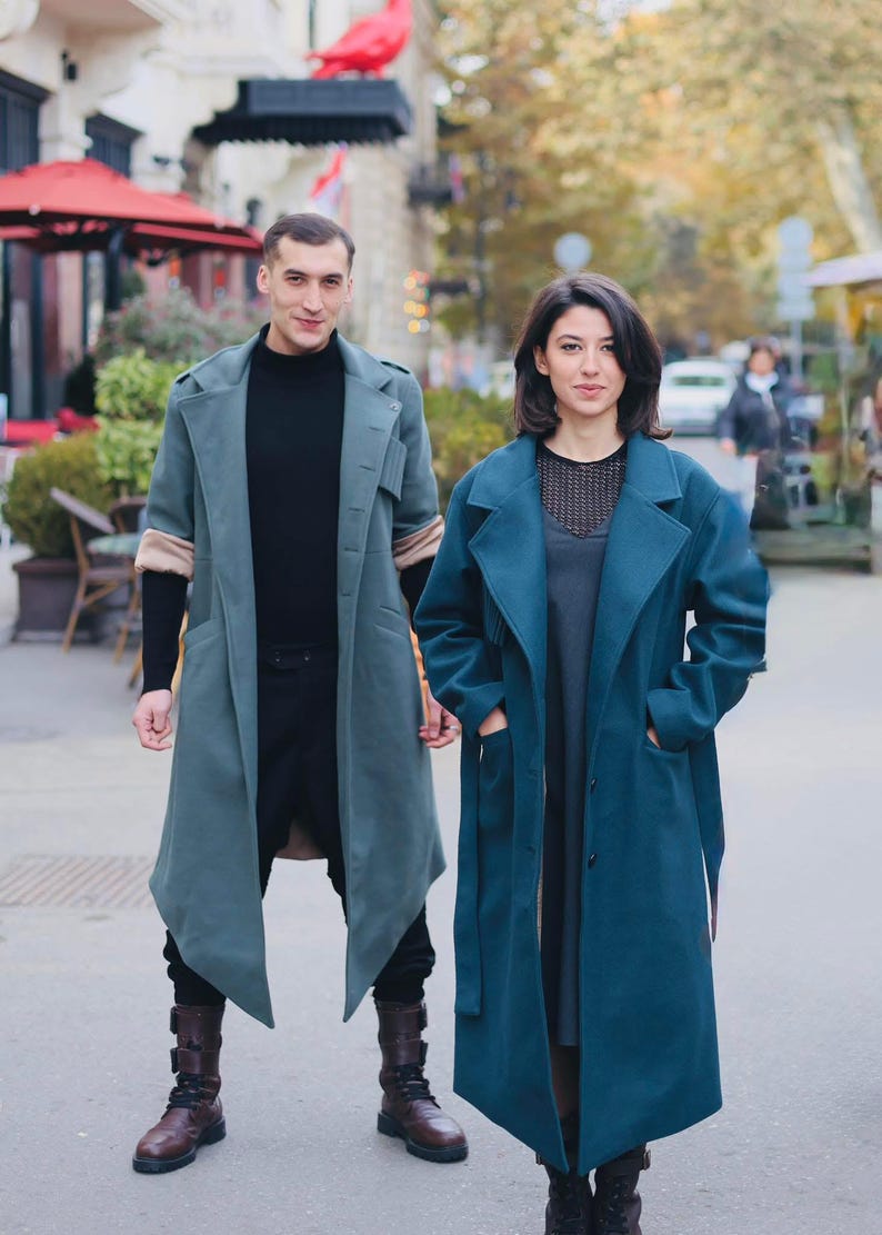 Op de afbeelding: Twee mensen in lange, teal-kleurige jassen. De jas links heeft beige mouwen en wordt gecombineerd met een zwarte broek en bruine laarzen. De jas rechts is donkerder teal en heeft een bijpassende riem. De achtergrond is een stadsstraat.