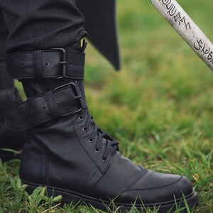 Op de afbeelding: Zwarte leren laarzen met veters en gespriemen. De laarzen worden gedragen met een zwarte broek en staan tegen een achtergrond van groen gras. De laarzen hebben een robuust, utilitair ontwerp.