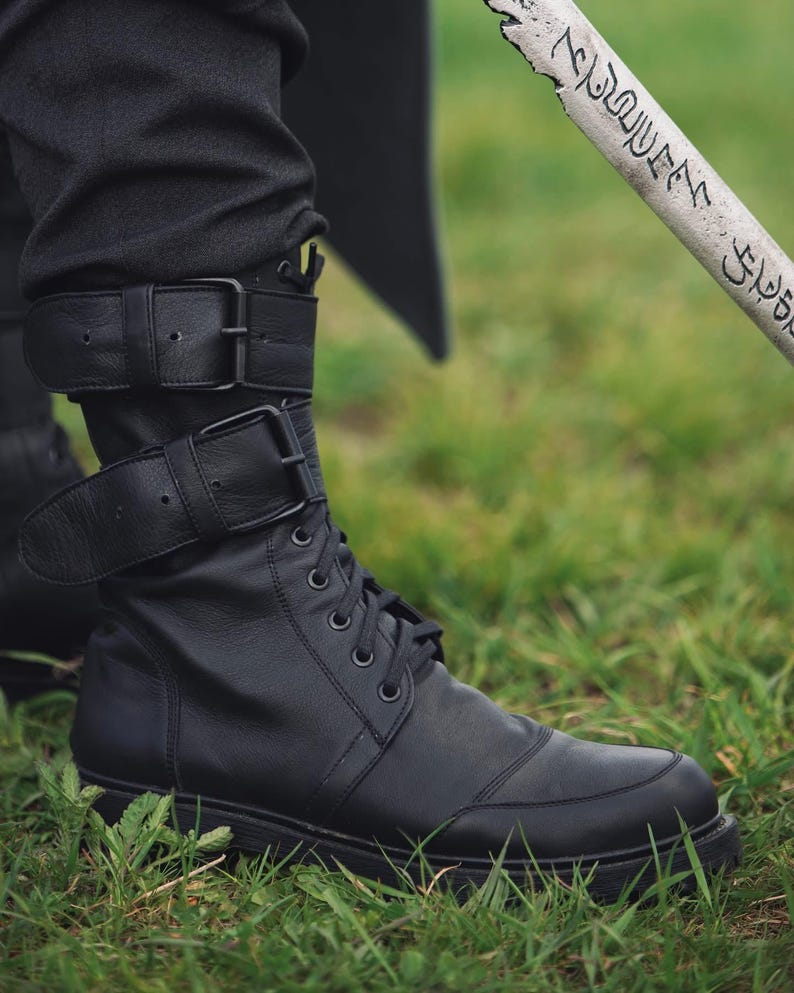 Op de afbeelding: Zwarte leren laarzen met veters en gespriemen. De laarzen worden gedragen met een zwarte broek en staan tegen een achtergrond van groen gras. De laarzen hebben een robuust, utilitair ontwerp.