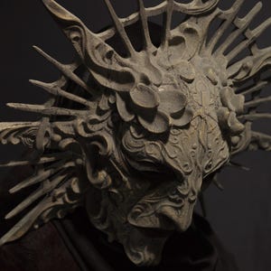 May include: A detailed, weathered mask with an intricate design. The mask is primarily gray with a textured surface and features ornate details, including floral and swirling patterns. Spikes extend from the top, creating a crown-like appearance. The mask is set against a dark background.