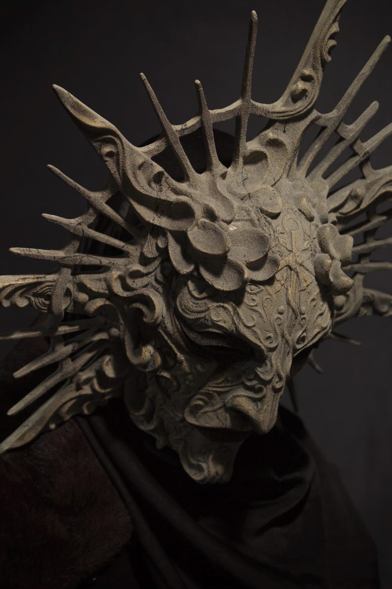 May include: A detailed, weathered mask with an intricate design. The mask is primarily gray with a textured surface and features ornate details, including floral and swirling patterns. Spikes extend from the top, creating a crown-like appearance. The mask is set against a dark background.