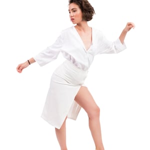 Puede incluir: Un atuendo blanco que incluye una camisa abotonada, una falda de cintura alta con abertura en el muslo y tacones con tiras. El conjunto completamente blanco crea un look limpio y elegante. La modelo está en una pose dinámica.