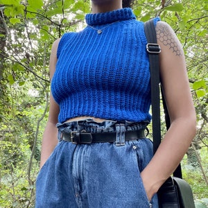 Peut inclure: Un haut bleu en crochet sans manches avec un col montant et un ourlet à volants. Le haut est porté avec un jean bleu et une ceinture en cuir noir.