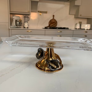 May include: A clear glass rectangular cake stand with a gold base featuring two black floral accents.