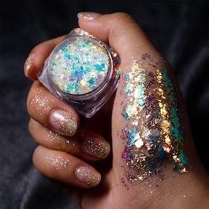 May include: A small, clear, square cosmetic jar filled with iridescent, multi-colored flakes that shift between blue, gold, and purple. The same reflective, metallic-looking flakes are applied to the skin, creating a shimmering, textured effect.