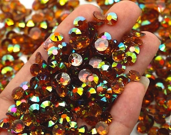 May include: A close-up of a hand holding a large quantity of iridescent amber-colored rhinestones. The rhinestones are faceted and have a rainbow-like sheen.