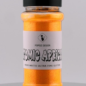 May include: A glass jar filled with bright orange neon matte ultra fine glitter. The jar has a black lid and a white label that reads "POPOZ DESIGN ATOMIC APRICOT NEON MATTE ULTRA FINE GLITTER".