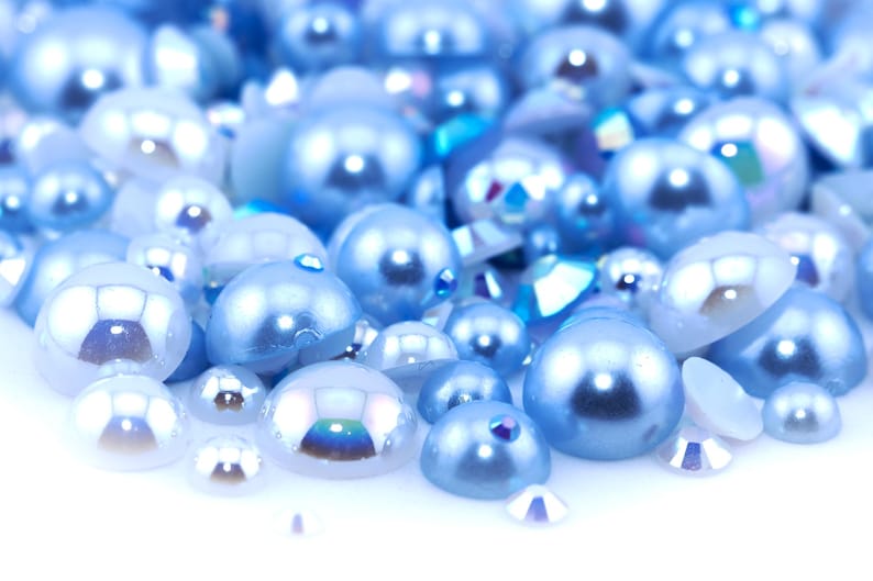 May include: A close-up of a pile of light blue iridescent pearls. The pearls are scattered on a white surface.