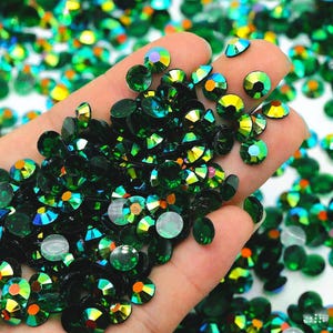 May include: A large quantity of green iridescent rhinestones, also known as glass gems, in various sizes. The rhinestones are scattered on a surface and a hand is holding a handful of the rhinestones.