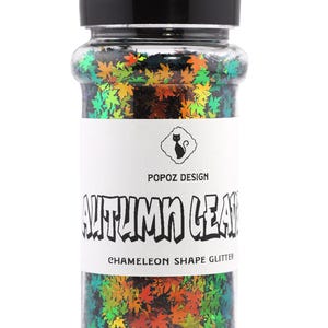 May include: A clear plastic jar with a black lid filled with autumn-themed glitter. The glitter is shaped like maple leaves in shades of green, orange, and yellow. The label reads "AUTUMN LEAF" and "CHAMELEON SHAPE GLITTER".