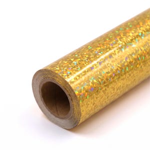 May include: A roll of gold-colored adhesive vinyl featuring a reflective holographic finish with a repeating geometric pattern that creates a shimmering, multi-colored effect across the surface of the material when light hits the metallic texture.