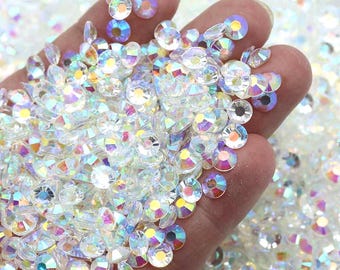 May include: A hand holding a large amount of iridescent clear glass rhinestones. The rhinestones are small and round, with a flat back and a faceted front. They are scattered on a white surface.