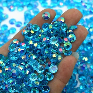 May include: A hand holding a large quantity of iridescent blue rhinestones. The rhinestones are round and have a flat back.
