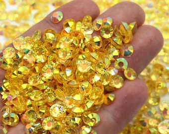 May include: A handful of sparkling, yellow-gold rhinestones. The faceted gems are round with a flat back and are scattered across the image. The gems have a bright, reflective quality, catching the light and creating a dazzling effect. The overall theme is craft supplies.