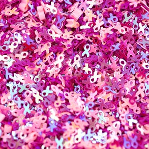 May include: Close-up of a vibrant assortment of pink ribbon-shaped confetti. The confetti pieces vary in shades of pink, from light to deep magenta, with some iridescent accents. The scattered shapes create a textured, celebratory appearance.