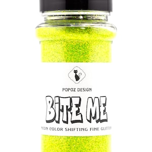 May include: A clear glass jar filled with neon green glitter. The jar has a black lid and a white label with the text "BITE ME" and "NEON COLOR SHIFTING FINE GLITTER". The label also features a black cat logo.