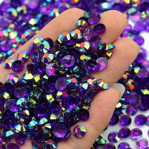 May include: A large quantity of iridescent purple rhinestones, with a rainbow effect, are scattered in a hand. The rhinestones are small and round, with a flat back and a pointed top.