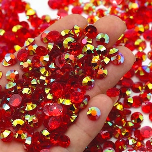 May include: A large pile of red iridescent round rhinestones. The rhinestones have a flat back and a faceted front. The rhinestones are scattered on a hand.
