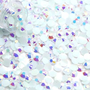 May include: A close-up of a large quantity of iridescent, light blue, flat-back rhinestones. The rhinestones are scattered on a white surface.