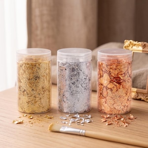 May include: Three clear cylindrical plastic jars filled with metallic foil flakes in gold, silver, and copper colors. Each jar has a translucent lid. A wooden-handled brush with light-colored bristles lies horizontally in front of the jars.