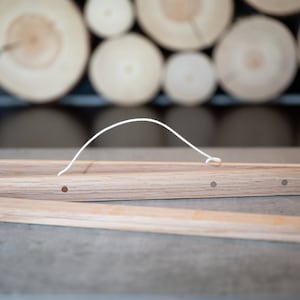May include: A wooden poster hanger with a white string handle. The hanger is made of two pieces of wood that are joined together with a small metal pin. The hanger has four holes in it, which are used to hang the poster.