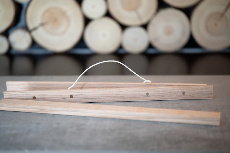 May include: A wooden poster hanger with a white string handle. The hanger is made of two pieces of wood that are joined together with a small metal pin. The hanger has four holes in it, which are used to hang the poster.