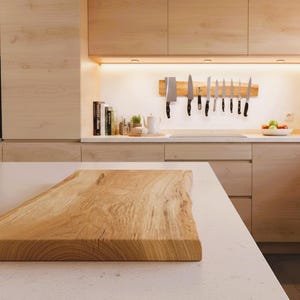 May include: A light-colored wooden cutting board with a natural edge sits on a white countertop in a modern kitchen. The kitchen features light wood cabinets, a black oven, and a knife rack with various knives.