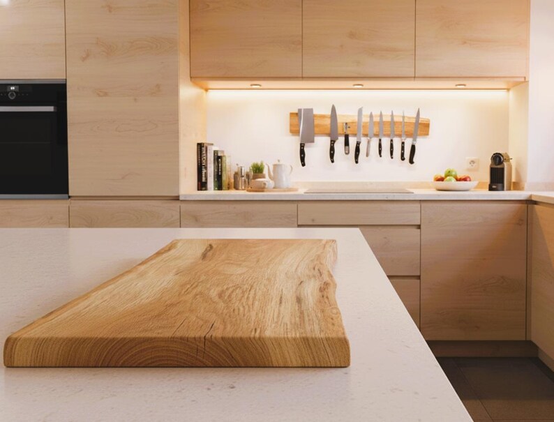 May include: A light-colored wooden cutting board with a natural edge sits on a white countertop in a modern kitchen. The kitchen features light wood cabinets, a black oven, and a knife rack with various knives.