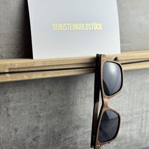Könnte beinhalten: Sonnenbrillenhalter aus Holz mit schwarzem Kordeldetail, der eine braune Sonnenbrille hält. Eine weiße Karte mit goldenem Text "DUBISTEINGOLDSTÜCK" befindet sich im Halter. Der Halter ist an einer grauen Wand montiert.
