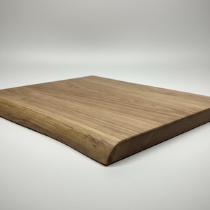 Puede incluir: Tabla de cortar cuadrada de madera marrón claro con un borde natural en un lado. La tabla tiene una superficie lisa y esquinas redondeadas. La veta de la madera es visible. Ideal para la preparación de alimentos.
