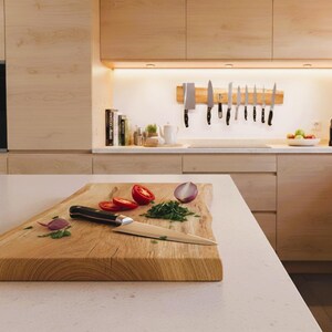 May include: A wooden cutting board with sliced tomatoes, chopped herbs, and a knife. A red onion is also visible. Kitchen knives are mounted on a wooden strip on the wall. The kitchen has light wood cabinets and a white countertop.