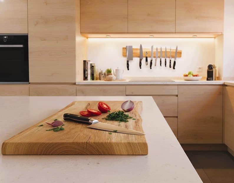 May include: A wooden cutting board with sliced tomatoes, chopped herbs, and a knife. A red onion is also visible. Kitchen knives are mounted on a wooden strip on the wall. The kitchen has light wood cabinets and a white countertop.