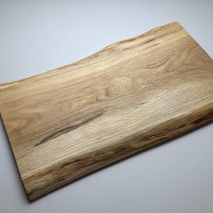 May include: A rectangular wooden cutting board with a natural, live edge. The board is made of light-colored wood with darker grain patterns and measures approximately 18 inches long and 12 inches wide.