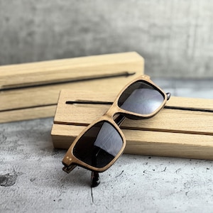 Könnte beinhalten: Eine Holzsonnenbrille mit dunklen Gläsern liegt auf einem hellen Holzständer. Die Sonnenbrille hat einen hellbraunen Rahmen mit dunklen Bügeln. Ein zweiter Holzständer befindet sich im Hintergrund. Die Ständer haben eine Nut.