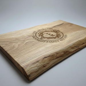 Può includere: Tagliere rettangolare in legno chiaro con bordo naturale. Il testo inciso recita "EST. 2024 - THE SCHMIDT FAMILY KITCHEN" attorno a un monogramma centrale "A&M".