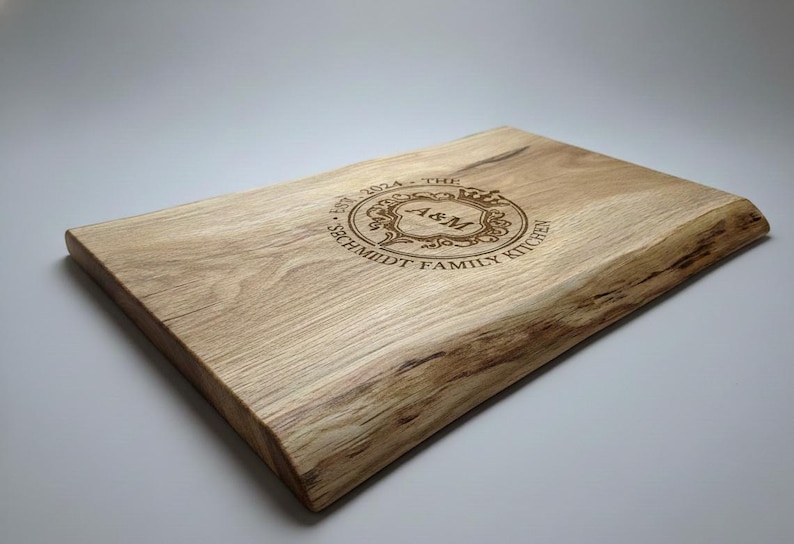 May include: A rectangular wooden cutting board with a natural edge. The board has a light brown color and features engraved text that reads "EST. 2024 - THE SCHMIDT FAMILY KITCHEN" around a central monogram "A&M".