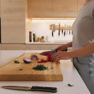 May include: A wooden cutting board with sliced red tomato, chopped green herbs, and a red onion half. A person is slicing a tomato with a black-handled knife. A second knife rests on the counter. Kitchen setting.