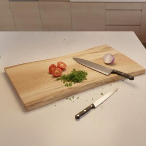 May include: A wooden cutting board with a large chef's knife and a smaller paring knife. Sliced tomatoes, chopped herbs, and a red onion half are arranged on the board. The kitchen setting has light-colored cabinets.