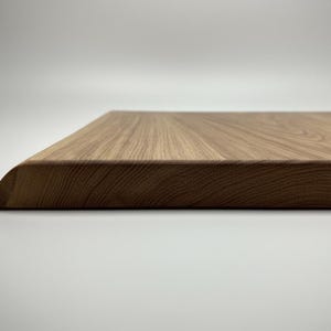 May include: Close-up of a rectangular wooden cutting board with a rounded corner. The board has a natural wood grain pattern and a light brown color. The board is likely used for food preparation.