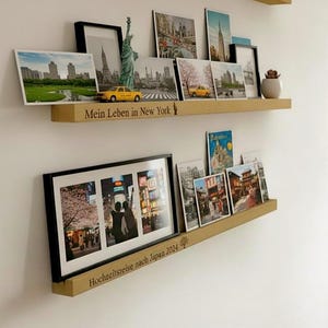 Op de afbeelding: Twee houten planken aan een witte muur, met ingelijste foto's, ansichtkaarten en kleine decoratieve items. De bovenste plank heeft de tekst "Mein Leben in New York". De onderste plank heeft de tekst "Hochzeitsreise nach Japan 2024".
